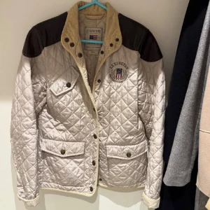 Beige quiltad jacka från Lexington - Quiltad beige jacka från Lexington med mörkbruna axelpartier och snygg broderad logga på bröstet. Jackan har flera fickor med knappar, hög krage i manchester och klassisk amerikansk outwear-vibe. Perfekt för dig som gillar stilren och sportig look.