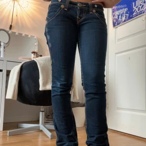 True Religion mörkblå jeans - Säljer ett par unika True Religion jeans i mörkblå tvätt med kontrastsömmar och snygga bakfickor med signaturbroderi. Modellen har låg midja och raka/bootcut ben lite beroende på person till person. Den har defekter längst ner på bakre benen  men förutom det är de i bra skick!