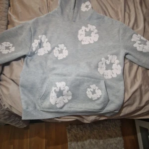 Grå denim tears hoodie - Säljer en grå hoodie med stora vita blommönster över hela tröjan. Hoodien har en stor ficka framtill och en skön huva. Perfekt för dig som gillar en chill och unik stil. Materialet känns mjukt och passar bra till vardags.