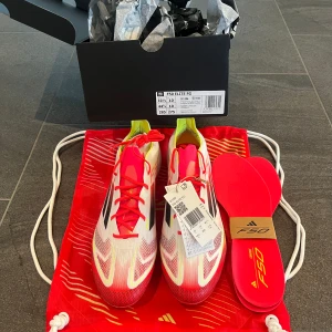 Adidas F50 Elite FG fotbollsskor - Säljer ett par Adidas F50 Elite FG fotbollsskor i vitt, rött och svart med gula detaljer. Skorna har snörning, låg profil och är tillverkade i lätt syntetmaterial för maxad speed på planen. Snygg design med svarta ränder och röda sulor. Perfekta för dig som vill sticka ut på matchen.