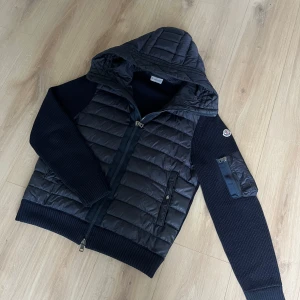 Moncler cardigan  - Snygg svart pufferjacka från Moncler med stickade ärmar och vadderad kropp. Jackan har huva, dragkedja framtill och en extra ficka med dragkedja på ärmen. Kvitto finns! Hör av dig om du har frågor! 