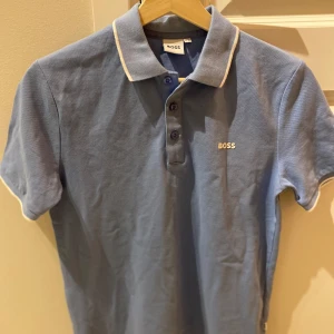 Ljusblå Piké Hugo Boss - Snygg blå pikétröja från BOSS med klassisk krage och tre knappar framtill. Tröjan har korta ärmar och vit kant på krage och ärmslut. Tillverkad i mjuk bomull och har BOSS-logga broderad på bröstet. Endast använd EN GÅNG! Storlek: 12-xs. Nypris 700kr. Hör av dig om du har några frågor elr liknande🙌