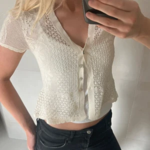 Vit blommig stickad blus från Zara - Supersöt vit stickad blus från Zara med korta ärmar och v-ringning. Blusen har fina blommiga detaljer i mönstret, knappar framtill och är lätt croppad. Perfekt till jeans eller kjol för en fräsch look.