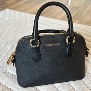Svart handväska från Michael Kors - Snygg svart handväska från Michael Kors med guldfärgade detaljer och dragkedja. Väskan har både handtag och en avtagbar axelrem med kedjedetaljer. Tillverkad i strukturerat skinn och har logga framtill. Perfekt storlek för det viktigaste du behöver ha med dig.