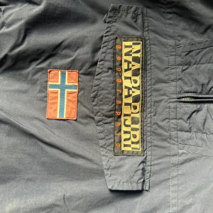 Blå vindjacka från Napapijri med huva - Säljer en blå vindjacka från Napapijri med stor logga och norsk flagga på bröstet. Jackan har huva, halv dragkedja vid sidan och funkar som en hoodie, och en stor ficka framtill. Perfekt för dig som gillar streetwear och vill ha en snygg och funktionell jacka.