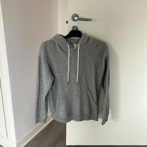 Grå hoodie med huva och ficka - En basic grå hoodie med vit snörning i huvan och en stor magficka. Tröjan har lång ärm och är gjord i mjuk bomullsmix, perfekt för chill dagar. Enkel och stilren design som funkar till det mesta.