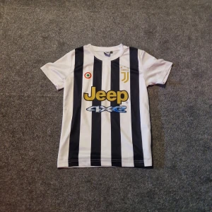 Juventus fotbollströja svartvit randig - Juventus matchtröja med klassiska svarta och vita ränder, Jeep-logga i guld och klubbmärke på bröstet. Kortärmad med rund hals och tillverkad i lätt polyester som andas. Perfekt för dig som vill representera ditt favoritlag på planen eller på läktaren.