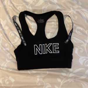 Svart sporttopp från Nike - Sportig svart topp från Nike med bred resår och vita loggor på axelbanden. Toppen har en racerback-design och är tillverkad i ett stretchigt polyestermaterial som passar perfekt för träning. Stort Nike-tryck framtill ger en cool look. Priset kan diskuteras 