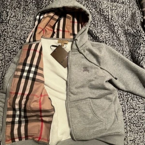 Grå Burberry hoodie med klassiskt foder - Snygg grå hoodie från Burberry med dragkedja och huva. Insidan av huvan har det ikoniska rutiga Burberry-mönstret i beige, svart, rött och vitt. Tröjan har två fickor fram och diskret broderad logga på bröstet. Perfekt för dig som vill ha en stilren och lyxig look.