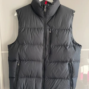 Polo Ralph Lauren väst - Snygg svart dunväst från Polo Ralph Lauren med hög krage och tre praktiska fickor med dragkedja. Västen har ett stilrent, quiltat mönster och diskret logga på bröstet. Perfekt att slänga över en hoodie när det är kyligt ute.