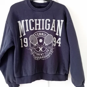 Sweatshirt Gina Tricot S - Cool mörkgrå sweatshirt från Gina Tricot med stort Michigan-tryck och tennismotiv framtill. Tröjan har rund hals, långa ärmar och ribbade muddar. Perfekt för en sportig och avslappnad stil.