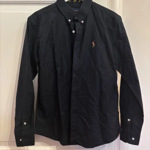 Mörkblå Ralph Lauren skjorta - Klassisk mörkblå skjorta från Ralph Lauren i bomull med broderad logga på bröstet. Skjortan har lång ärm, knappar framtill och button down-krage. Custom fit-modell som sitter snyggt och passar till många olika stilar. Skick 10/10. Är storlek M men passar även S. Fråga på!