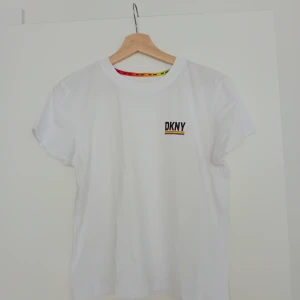 Vit DKNY t-shirt XS  - Vit t-shirt från DKNY Sport med korta ärmar och rund hals. På bröstet finns en svart DKNY-logga med en färgglad regnbågsrand under. Halsringningen har en insida med DKNY Sport-tryck i rött, orange och gult. Tillverkad i mjuk bomull. I toppskick, som ny!