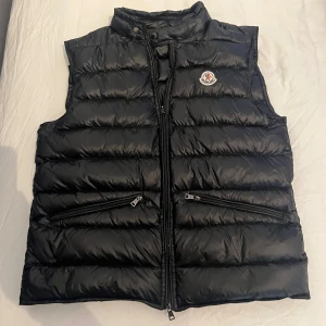Svart dunväst från Moncler - Snygg svart dunväst från Moncler med klassisk quiltad design och hög krage. Moncler väst gui. Storlek 2 vilket motsvarar ungefär S. Mitt pris 1400 priset kan diskuteras. Skick 10/10 inga defekter. Alla nfc taggar funkar och leder dig direkt till monclers hemsida. Vid frågor/intresse/bilder skriv!