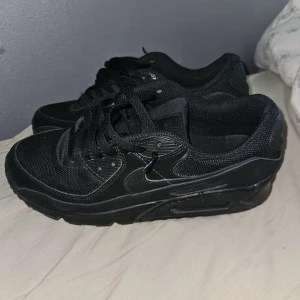 Svarta Nike Air Max sneakers - Säljer ett par helsvarta Nike Air Max sneakers med klassisk chunky sula och meshdetaljer. Skorna har snörning och är tillverkade i en mix av syntet och textil. Perfekta för dig som gillar en clean och sportig look.