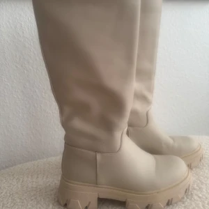 Beiga chunky boots i skinnimitation - Säljer ett par höga boots i beige skinnimitation med chunky sula och rund tå. Snyggt minimalistisk design utan snörning, perfekt till jeans eller kjol. Storlek 36 och riktigt trendiga för höst och vinter. Använd en gång.
