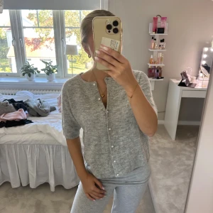 Grå kofta med kort ärm och pärlknappar - Superfin ljusgrå kofta med korta ärmar och pärlknappar framtill. Koftan är stickad och har en rund halsringning, perfekt att slänga på sig över ett linne eller t-shirt. Enkel och stilren design som passar till många outfits.