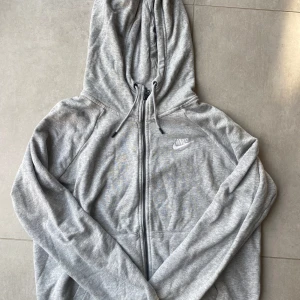 Grå hoodie från Nike med dragkedja - Säljer en klassisk grå hoodie från Nike med broderad logga på bröstet. Tröjan har huva med snörning, hel dragkedja och långa ärmar. Perfekt för chill dagar eller när du vill ha en sportig look. Ingen storlek står men uppskattas till M.