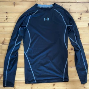 Svart långärmad träningströja Under Armour - Svart långärmad kompressionströja från Under Armour med gråa sömmar och logga på bröstet. Tillverkad i syntetmaterial, perfekt för träning. Tight passform och HeatGear-teknologi som håller dig sval under intensiva pass.