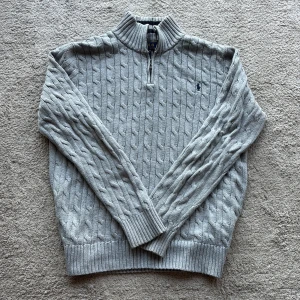 Grå kabelstickad tröja Polo Ralph Lauren - Kabelstickad grå tröja från Polo Ralph Lauren med halv dragkedja och ribbad krage med mörkblå ränder. Klassisk logga broderad på bröstet och långa ärmar. Perfekt för lager-på-lager och chill höst- eller vinterstil.