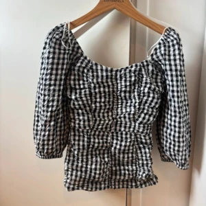 Rutig blus med puffärm från Bik Bok - Supersöt svartvit rutig blus från Bik Bok med smockad front och rygg. Blusen har puffiga långärmar med resår vid ärmslut och en fyrkantig urringning. Perfekt att styla med jeans eller kjol för en trendig look.