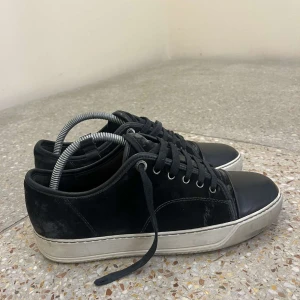 Svarta sneakers från Lanvin i mocka - Snygga svarta sneakers från Lanvin med ovandel i mocka och tåparti i svart skinn. Skorna har vita, rejäla gummisulor och klassisk snörning. Perfekta för dig som gillar stilrena och lyxiga sneakers. Skorna har minimala skador (bild 5), annars är skicket hyfsat bra. OBS! Box ingår inte men dustbag ingår. Kan gå ner i pris vid snabbaffär. Hör av er vid minsta lilla fråga