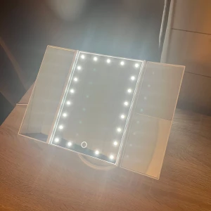 Sminkspegel med LED-belysning - Snygg sminkspegel i vit plast med tre paneler och inbyggd LED-belysning runt mittenspegeln. Touchknapp på framsidan för att tända/släcka lamporna. Spegeln står stadigt på en oval fot och har vikbara sidor för bättre vinkel och närbild. Man kan ha lyset igång me både sladd som du får me och du kan även använda batteri