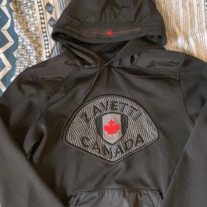 Svart hoodie från ZAVETTI Canada - Svart hoodie från ZAVETTI Canada med stor broderad logga och röd lönnlöv på bröstet. Tröjan har huva med snörning och lång ärm, tillverkad i mjukt syntetmaterial. Perfekt för dig som gillar streetwear och vill sticka ut med en unik design. Mycket bra skick. Pris kan diskuteras vid snabb affär