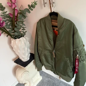 Alpha Industries bomberjacka, grön & vinröd - Säljer två klassiska bomberjackor från Alpha Industries, en i olivgrönt och en i vinrött. Jackorna har orange foder, ribbade muddar och ikonisk zip-puller med 'Remove Before Flight'. Perfekt för dig som vill ha en streetig och tidlös look. Den vin röda är storlek L och den gröna är M
