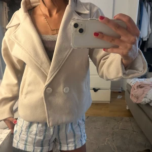 Beige kappa med knappar - Snygg beige kappa med bred krage och två stora vita knappar framtill. Jackan är kortare i modellen och har en stilren, minimalistisk look. Perfekt för dig som gillar en clean och trendig vibe. Fodrad insida för extra komfort.