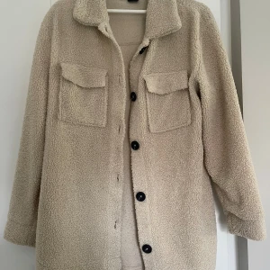 Beige teddyjacka från Gina Tricot - Supermjuk och oversized teddyjacka i beige från Gina Tricot. Jackan har stora bröstfickor med lock, svarta knappar framtill och klassisk krage💛💛💛