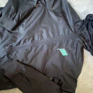 Hammarby jacka  - Svart vindjacka i storlek S med dragkedja och huva. Hammarby jacka , ribbade muddar. Perfekt för blåsiga dagar och enkel att matcha med streetwear. Jackan har ett litet hål i jackfickan men enkelt att sy ihop. Aven två små hål som du kan se på 4e bilden. Finns ej att köpa längre 