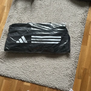 Svart sportväska från Adidas - Oöppnad!!Säljer en svart sportväska från Adidas med klassiska vita ränder och logga på sidan. Perfekt storlek för gymmet eller träningen, med praktisk form och tillverkad i slitstarkt syntetmaterial. Väskan är enkel att bära och har en stilren look.