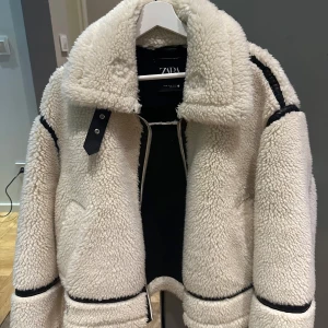 Vit teddyjacka från Zara med svarta detaljer - Säljer en fluffig och oversized vit teddyjacka från Zara med svarta skinnliknande detaljer längs sömmarna och vid kragen. Jackan har dragkedja framtill, stor krage och coolt bälte vid halsen. Perfekt för kalla dagar när du vill vara både varm och snygg. Har bara använts en gång har inga skador eller slitage original pris 1195kr 