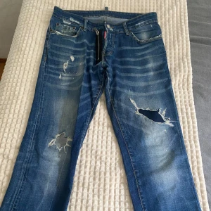 Blå slitna jeans med hål och detaljer - Säljer ett par blå jeans med slitningar, hål på båda benen och coola slitna detaljer. Jeansen har klassisk femficksdesign, dragkedja och knapp. Färgen är mellanblå med ljusare partier och lite färgstänk för en extra edge.