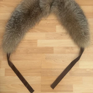 Brun/grå fluffig pälshalsduk med band - Supermjuk och fluffig halsduk/krage i brun och grå päls med långa bruna band att knyta med. Perfekt för att ge din outfit en lyxig touch och hålla dig varm under kalla dagar. Insidan är quiltad för extra komfort. Med foder.