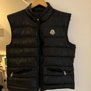 Svart dunväst från Moncler - Snygg svart dunväst från Moncler med quiltad design och hög krage. Västen har två fickor med dragkedja och klassisk Moncler-logga på bröstet. Perfekt för lager-på-lager och chill stil. Stängs med dragkedja och tryckknapp upptill.