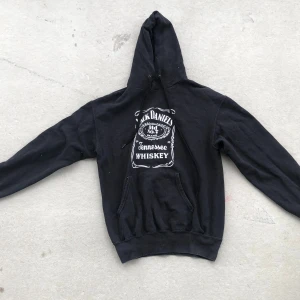Jack Daniel's svart hoodie med tryck - Svart hoodie med stor vit Jack Daniel's Tennessee Whiskey-tryck både fram och bak. Klassisk känguruficka, snörning i huvan och ribbade muddar. Perfekt för dig som gillar statement-plagg och vill sticka ut med en ikonisk look.