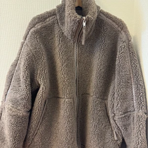 Beige teddyjacka med dragkedja - Mysig och oversized teddyjacka i beige med hög krage och hel dragkedja framtill. Jackan har två fickor och är tillverkad i fluffigt teddy-material som håller dig varm under kyliga dagar. Perfekt för dig som gillar en chill och trendig stil.