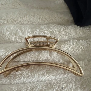 Guldig hårklämma i metall - Stilren hårklämma i guldigt metall med öppen design och rejäla taggar som håller håret på plats. Perfekt accessoar för att snabbt fixa en snygg frisyr. Klämman har en bågformad siluett och ger en trendig touch till din look.