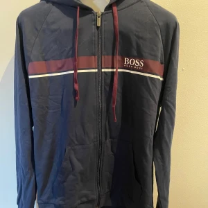 Marinblå hoodie från Hugo Boss - Snygg marinblå hoodie från Hugo Boss med dragkedja och huva. Tröjan har röda snören, en röd och vit rand över bröstet samt logga på vänster sida. Tillverkad i mjuk bomull och har praktiska fickor framtill. Perfekt för en chill och stilren look.