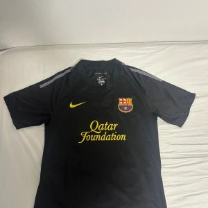 FC Barcelona svart Nike fotbollströja M - Snygg svart FC Barcelona fotbollströja från Nike med gult klubbmärke och Qatar Foundation-tryck på bröstet. Tillverkad i Dri-Fit-material som andas, perfekt för träning eller match. Klassisk passform med korta ärmar och rund hals.  Away kit 2011/12 Barcelona 