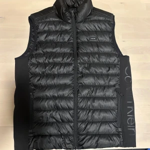HELT NY Svart dunväst från Calvin Klein - Snygg svart dunväst från Calvin Klein med quiltad design och hög krage. Västen har dragkedja framtill, två sidofickor med dragkedja och ett stort Calvin Klein-tryck längs sidan. Tillverkad av 100% återvunnen polyester, perfekt för lager-på-lager under kyliga dagar.