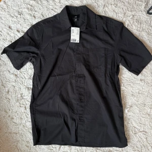 Svart kortärmad skjorta H&M XS - Svart skjorta från H&M i avslappnad passform med korta ärmar. Helt ny aldrig använd