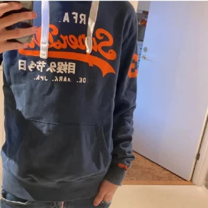 Blå Superdry hoodie med tryck - Säljer en blå hoodie från Superdry med orange och vitt tryck framtill och på ärmen. Tröjan har huva med vita snören och en stor magficka. Perfekt för dig som gillar streetwear och vill ha en skön och snygg look. Ganska mycket av texten har åkt av!!!!! Hör av er vid intresse 