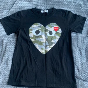 Svart Comme des Garçons Play t-shirt - Svart t-shirt från Comme des Garçons Play med stort hjärta i camouflageprint och ögon på bröstet. Riktigt fet och eftertraktad T-shirt. Säljer för 799, kan tänka mig att gå ner lite vid snabb affär. Tveka inte att höra av er med funderingar!