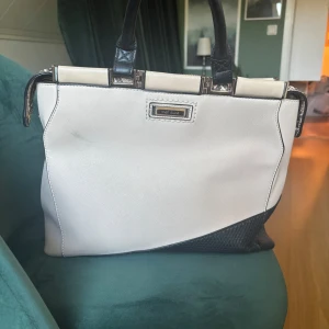 Vit och svart handväska från River Island - Snygg handväska från River Island i vit och svart med ormskinnsdetaljer och guldfärgade metalldetaljer. Väskan har två handtag, dragkedja bak och strukturerad botten i svart. Perfekt för dig som gillar stilrena accessoarer med en twist.