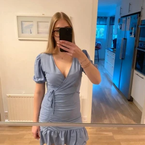 Ljusblå omlottklänning Gina Tricot - Supersöt ljusblå kort omlottklänning från Gina Tricot med puffärmar, v-ringning och volangkant nertill. Klänningen har knyt i midjan och är perfekt för sommarens alla tillfällen. Använd få gånger