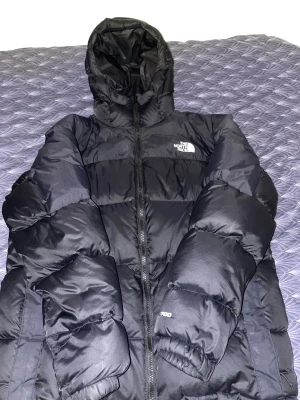 Svart Pufferjacka,  The North Face - Säljer en svart pufferjacka från The North Face med huva och dragkedja. Jackan är rejält vadderad och har logga på bröstet och axeln. Perfekt för kalla dagar och har en klassisk, stilren look. Passformen är normal och materialet är syntet med dunfyllning.