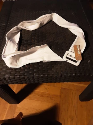 Vitt skärp från Miss Match - Snyggt vitt bälte från Miss Match med bred design och fyrkantigt spänne i samma färg. Bältet är gjort i skinnimitation och passar perfekt för att ge outfiten en clean och modern touch.106 lång